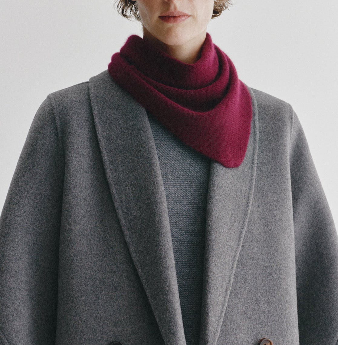 A person wearing a grey coat and sweater, styled with the Lisa Yang Florianne Scarf in burgundy draped around their neck, stands against a plain light background.