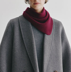 A person wearing a grey coat and sweater, styled with the Lisa Yang Florianne Scarf in burgundy draped around their neck, stands against a plain light background.