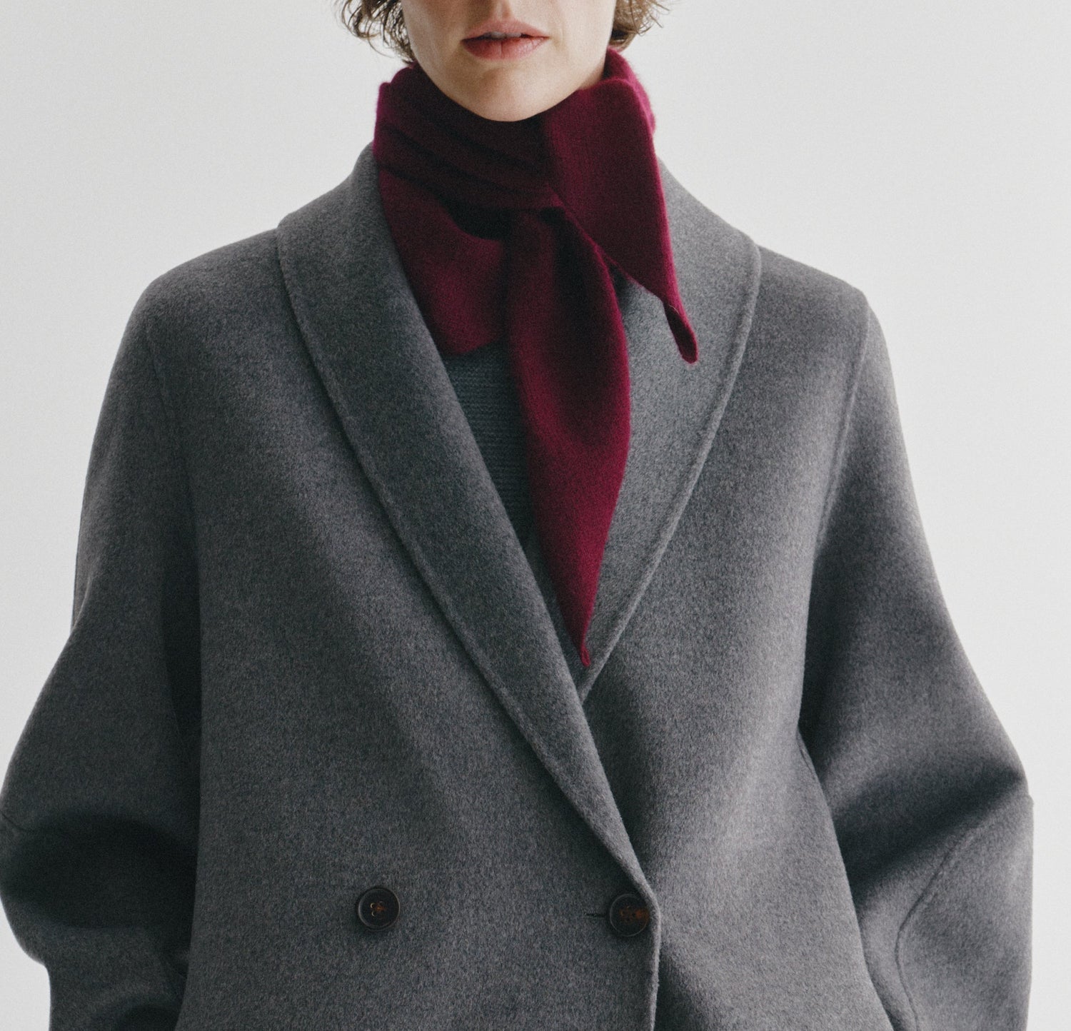 A person wearing a grey wool coat and the Lisa Yang Florianne Scarf in burgundy, tied at the neck, stands against a plain white background.
