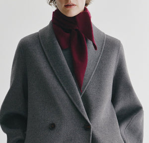 A person wearing a grey wool coat and the Lisa Yang Florianne Scarf in burgundy, tied at the neck, stands against a plain white background.