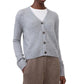 Wearing the Marionette Cardigan by Lisa Yang in light gray over a white shirt and brown high-waisted pants, the person stands with one hand in their pocket.