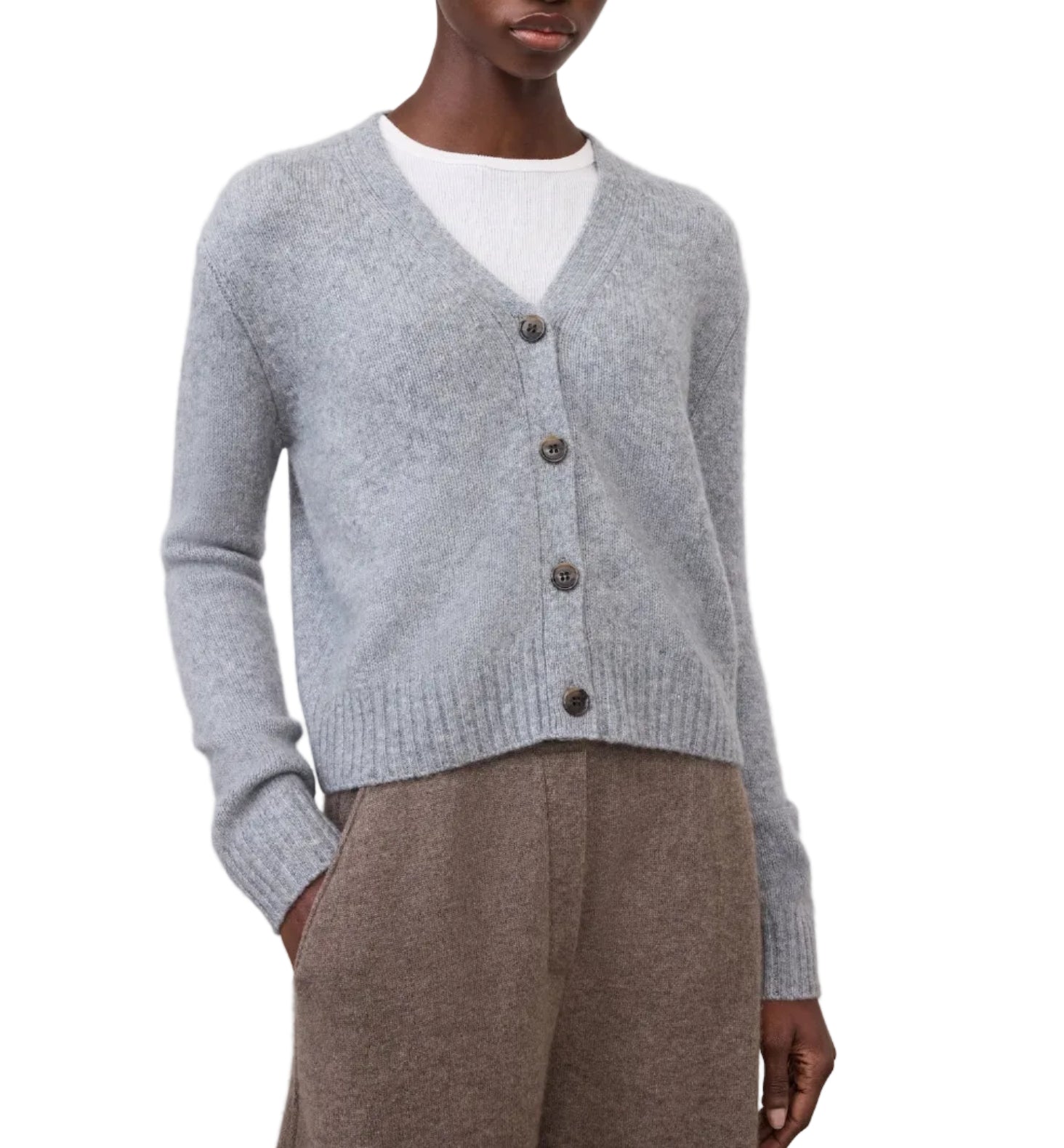 Wearing the Marionette Cardigan by Lisa Yang in light gray over a white shirt and brown high-waisted pants, the person stands with one hand in their pocket.