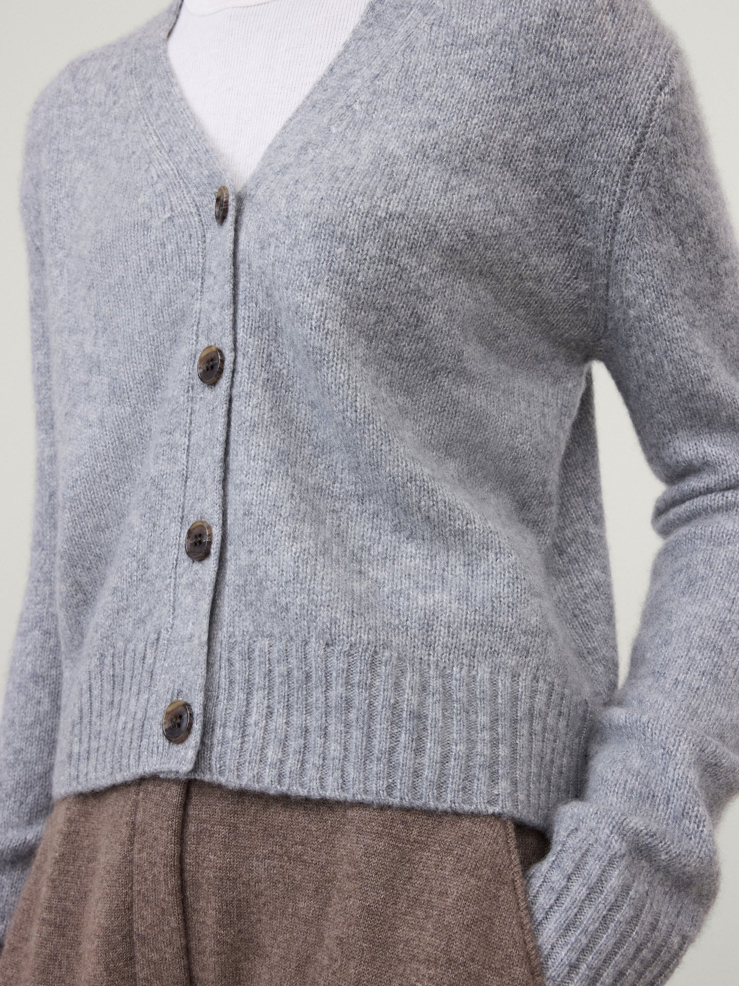 A person is wearing the Marionette Cardigan by Lisa Yang, a lightweight, light gray buttoned cardigan, over a white top and paired with high-waisted brown pants.