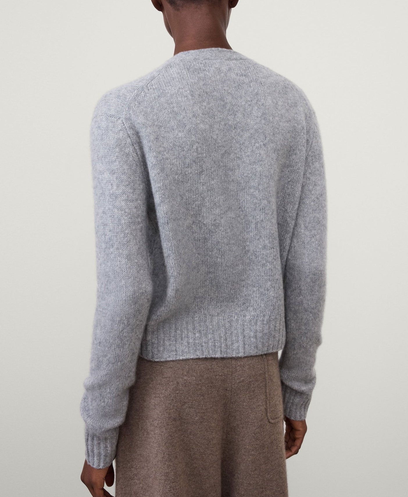 Model wearing the Lisa Yang Marionette Cardigan in light gray, paired with brown pants, shown from the back against a plain light background.