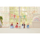 Four Calico Critters Biscuit Bear Family figurines, dressed in colorful outfits, stand on a white surface alongside a small toy house and book. They are set in front of a window decorated with rainbow bunting and soft pastel accents—ideal for sparking imaginative play or pairing with an architect toy set.