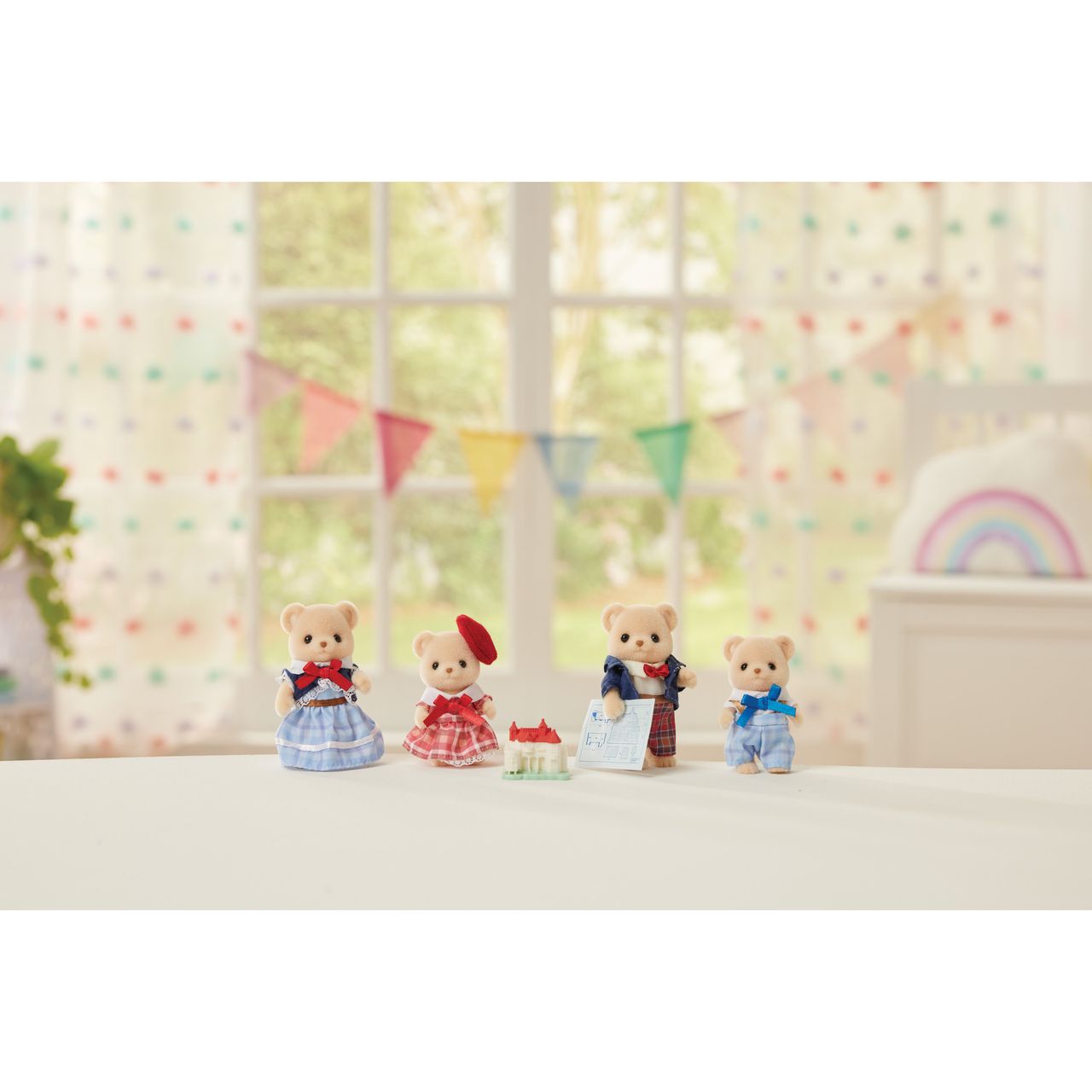 Four Calico Critters Biscuit Bear Family figurines, dressed in colorful outfits, stand on a white surface alongside a small toy house and book. They are set in front of a window decorated with rainbow bunting and soft pastel accents—ideal for sparking imaginative play or pairing with an architect toy set.