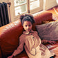 A young girl with curly hair sits on a brown leather couch, wearing the Louis Louise Girls' Belinda Big Corduroy Dress with heart-shaped pockets. A window and radiator are in the background.