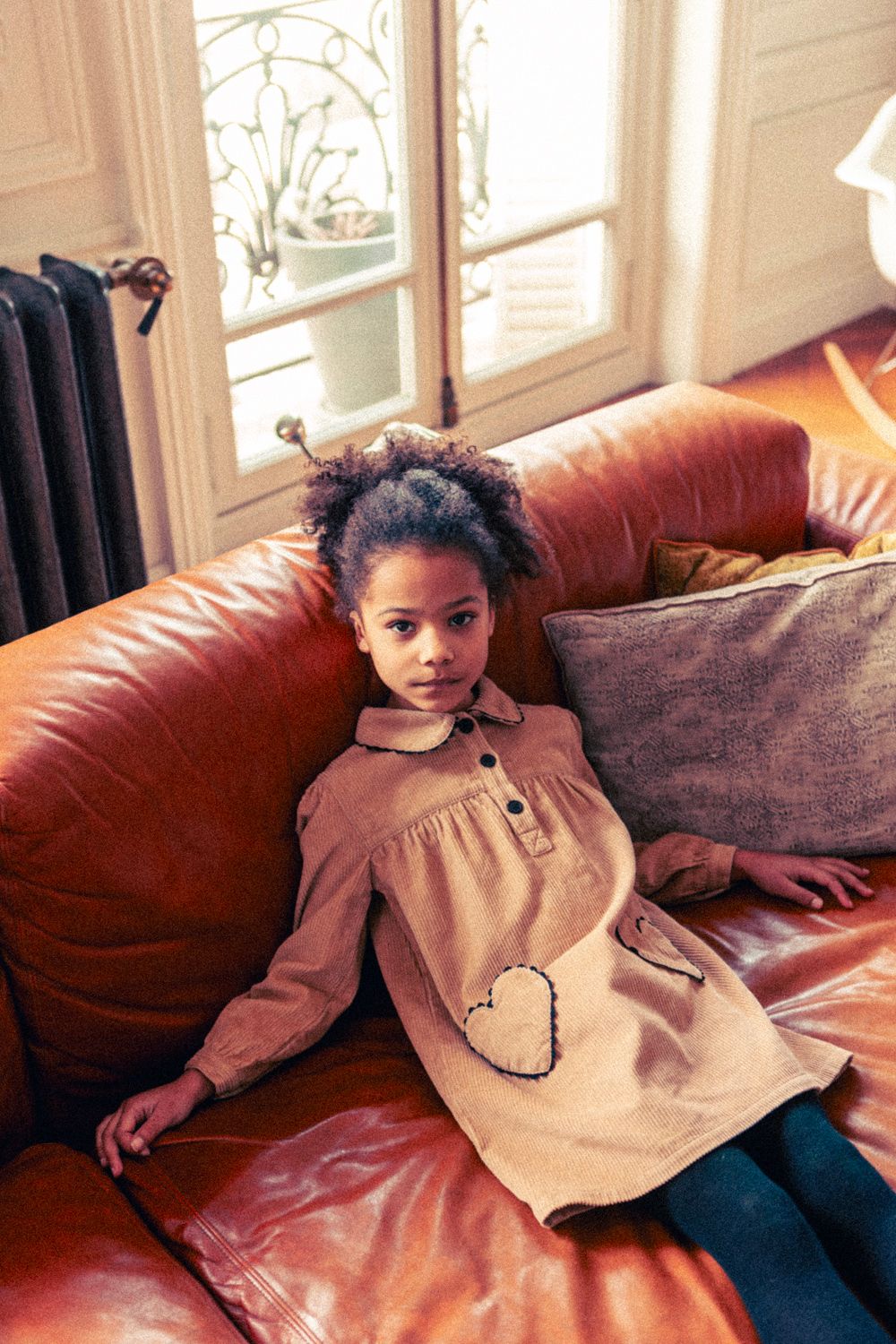 A young girl with curly hair sits on a brown leather couch, wearing the Louis Louise Girls' Belinda Big Corduroy Dress with heart-shaped pockets. A window and radiator are in the background.