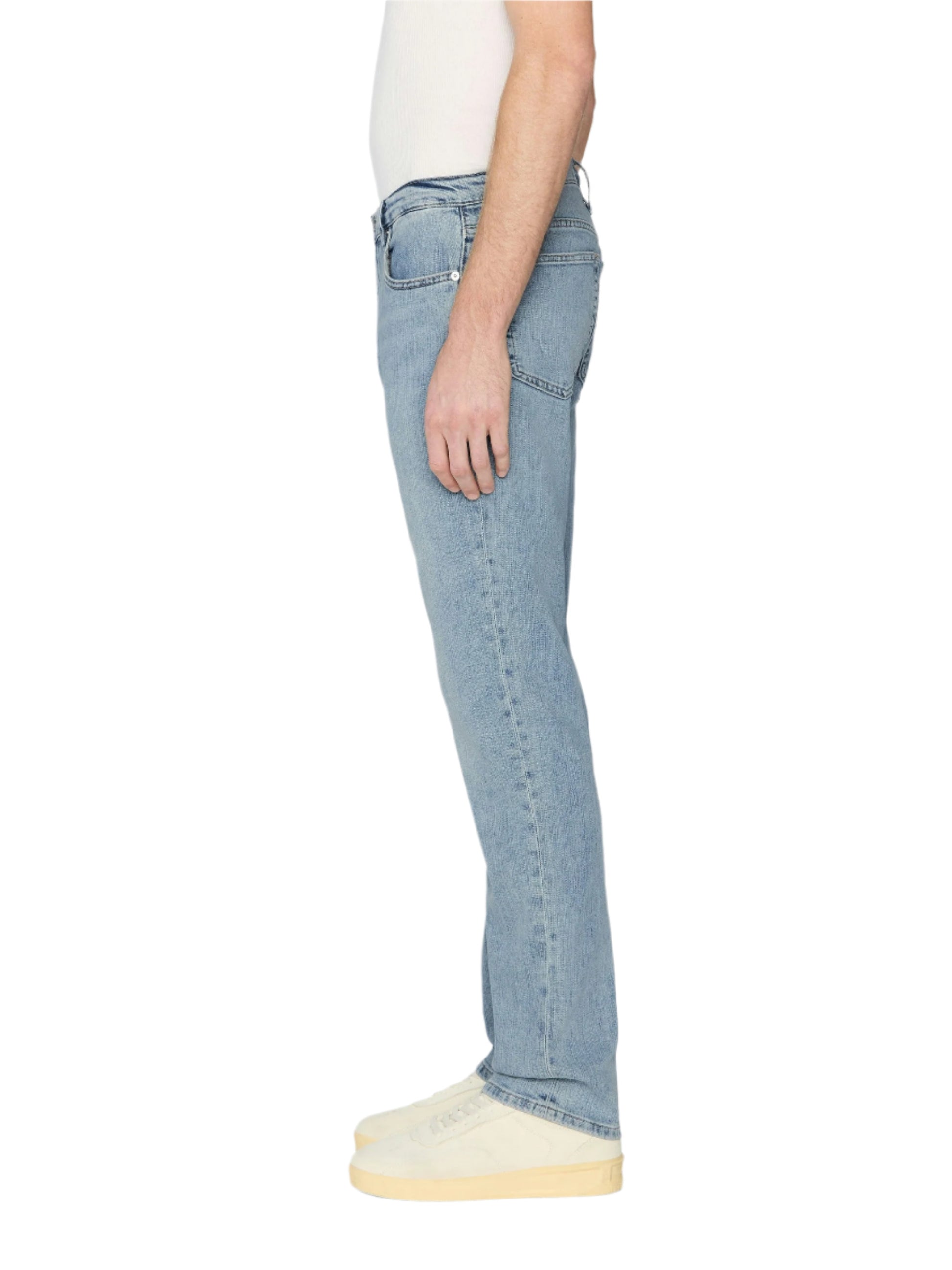 A person stands in side profile against a plain white background, wearing a white shirt, Frame L’Homme Slim Denim in light blue wash, and white shoes.
