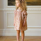 A young girl with curly hair wears the Noralee Girls' Maddie Dress, featuring vintage-inspired floral embroidery, paired with a matching bow and light shoes, as she stands indoors looking to the side.