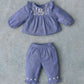 The Baby Girls’ Mikani Set by Louise Misha features a charming blue long-sleeve top adorned with white floral embroidery and delicate ruffles, paired with matching blue pants detailed with coordinating ruffle and flower accents. This beautifully embroidered blouse and pants set is showcased flat on a gray textured surface.