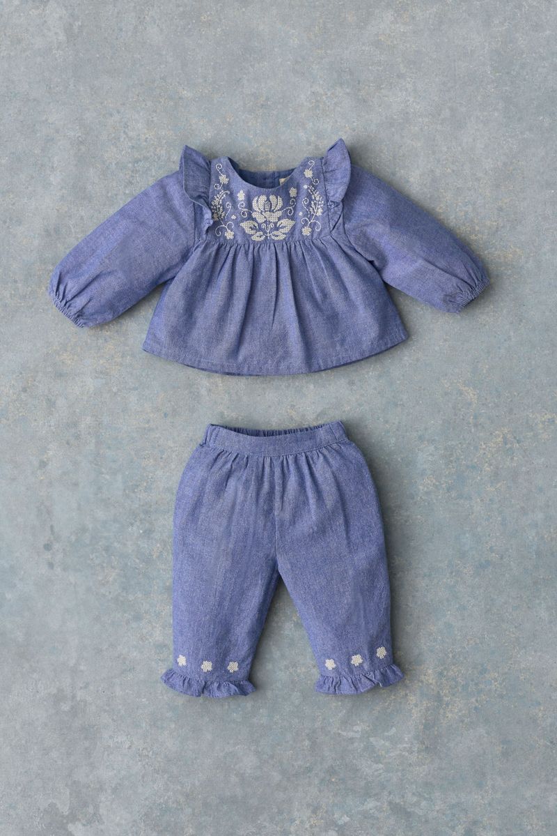 The Baby Girls’ Mikani Set by Louise Misha features a charming blue long-sleeve top adorned with white floral embroidery and delicate ruffles, paired with matching blue pants detailed with coordinating ruffle and flower accents. This beautifully embroidered blouse and pants set is showcased flat on a gray textured surface.