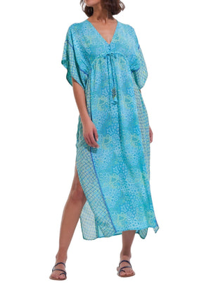 A person wearing the Poupette St Barth Amaya Kaftan, featuring a blue floral pattern with a flowy design, short sleeves, and a v-neck, stands with one leg slightly forward in sandals.