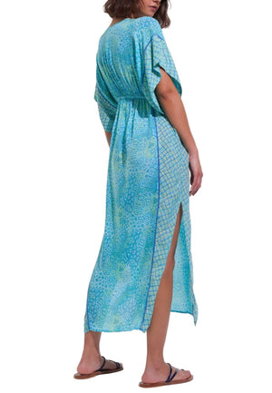 A person is seen from behind wearing the Poupette St Barth Amaya Kaftan, a blue patterned maxi dress adorned with vibrant floral motifs, equipped with short sleeves and a chic side slit.