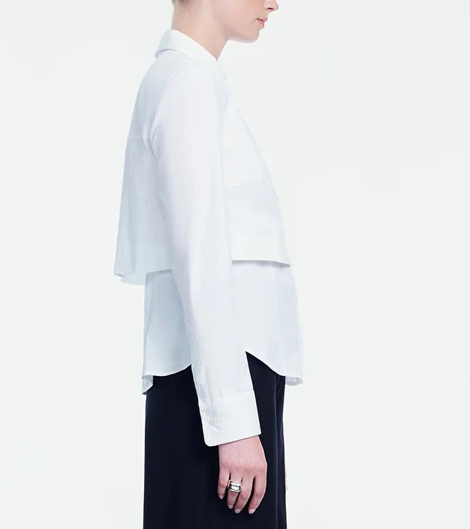 A person stands in profile against a plain white background, wearing the Lela Rose Cotton Poplin Layered Shirt in white with black pants.