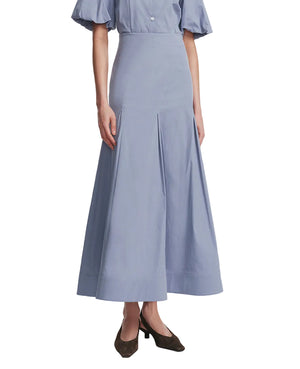 A woman wears the Lela Rose Seam Detail Skirt—a light blue, high-waisted, ankle-length A-line in soft cotton poplin with pleats—paired with a matching blouse and dark heels.