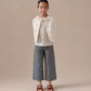 A young girl stands facing forward against a plain beige background, wearing a cream-colored jacket, patterned blouse, Bonpoint Girls Jingle Striped Denim Trousers, and brown sandals.