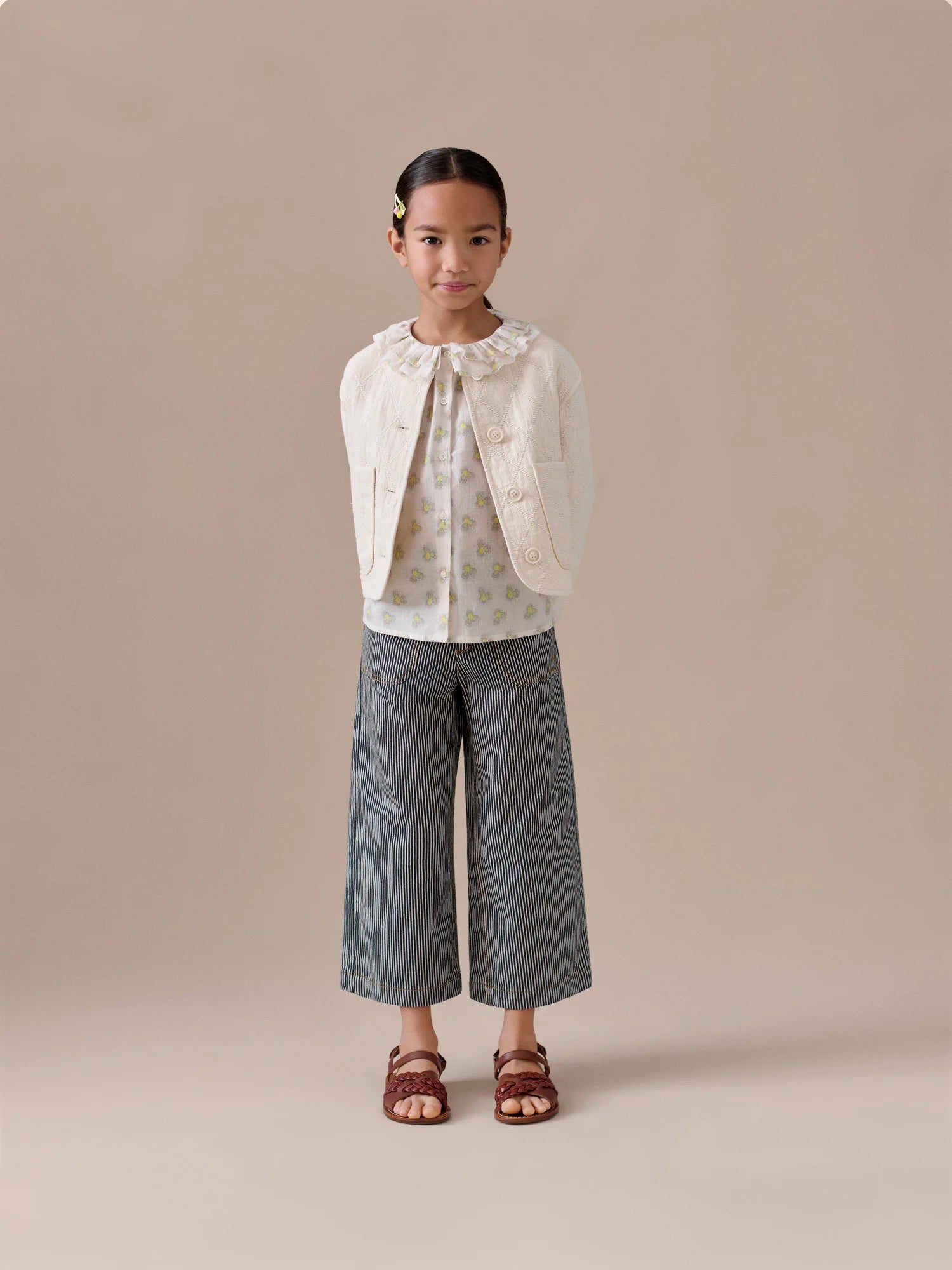 A young girl stands facing forward against a plain beige background, wearing a cream-colored jacket, patterned blouse, Bonpoint Girls Jingle Striped Denim Trousers, and brown sandals.