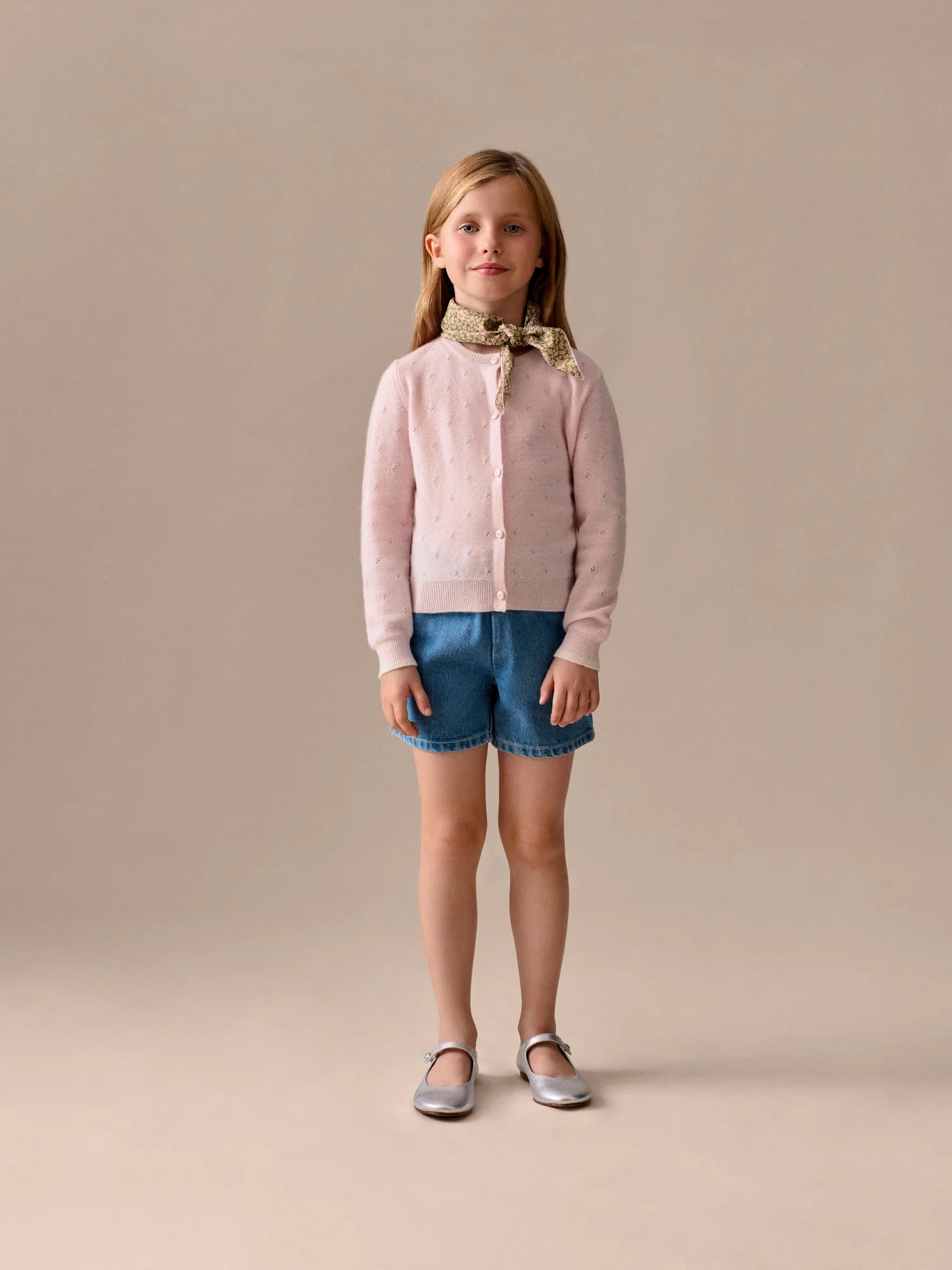 A young girl stands against a plain background in a pale pink cardigan, Bonpoint Girls' Milly Shorts with cherry embroidery, a patterned scarf, and silver shoes.