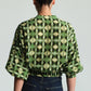 A person with dark hair in a bun is shown from the back wearing the Cara Cara Perrin Top—a green geometric-patterned top featuring wide sleeves and a cropped hem—paired with dark blue jeans.