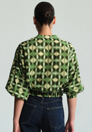 A person with dark hair in a bun is shown from the back wearing the Cara Cara Perrin Top—a green geometric-patterned top featuring wide sleeves and a cropped hem—paired with dark blue jeans.