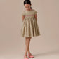 A young girl stands with hands behind her back, wearing the Bonpoint Girls' Duchesse Smocked Dress and pink shoes, looking down against a neutral background.
