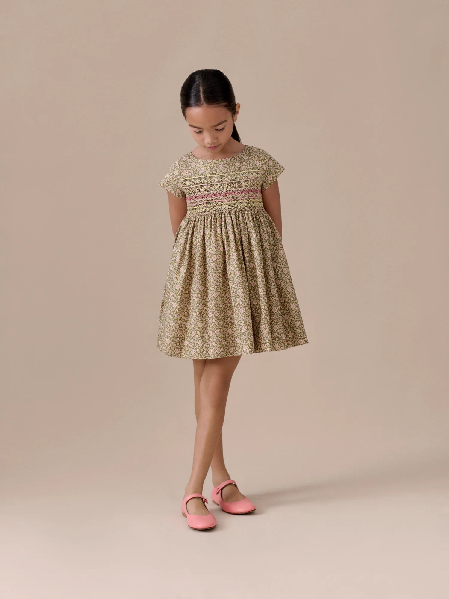 A young girl stands with hands behind her back, wearing the Bonpoint Girls' Duchesse Smocked Dress and pink shoes, looking down against a neutral background.