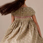 A girl with long brown hair wears the Bonpoint Girls' Duchesse Smocked Dress in green floral Liberty fabric, showing off its back buttons as her hair swings aside.