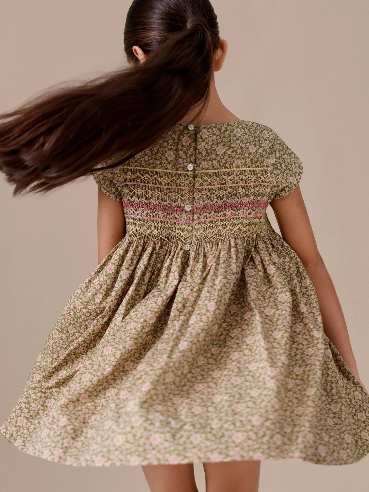 A girl with long brown hair wears the Bonpoint Girls' Duchesse Smocked Dress in green floral Liberty fabric, showing off its back buttons as her hair swings aside.
