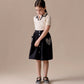 A young girl stands against a neutral background, wearing a short-sleeve cream top and the Bonpoint Girls' Lierre Hand-Embroidered Skirt with heart details on the pockets, looking down with her hands clasped.