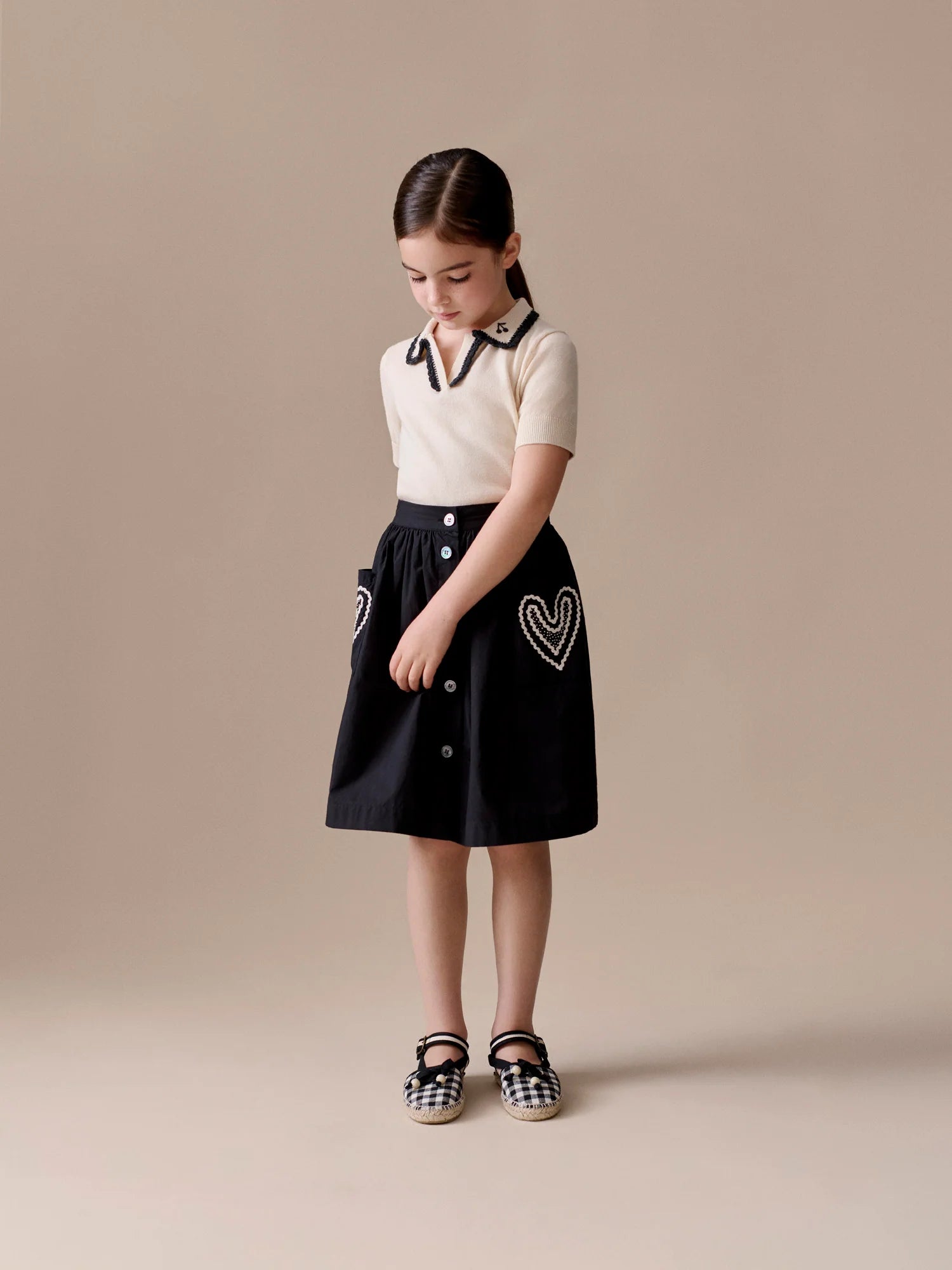 A young girl stands against a neutral background, wearing a short-sleeve cream top and the Bonpoint Girls' Lierre Hand-Embroidered Skirt with heart details on the pockets, looking down with her hands clasped.