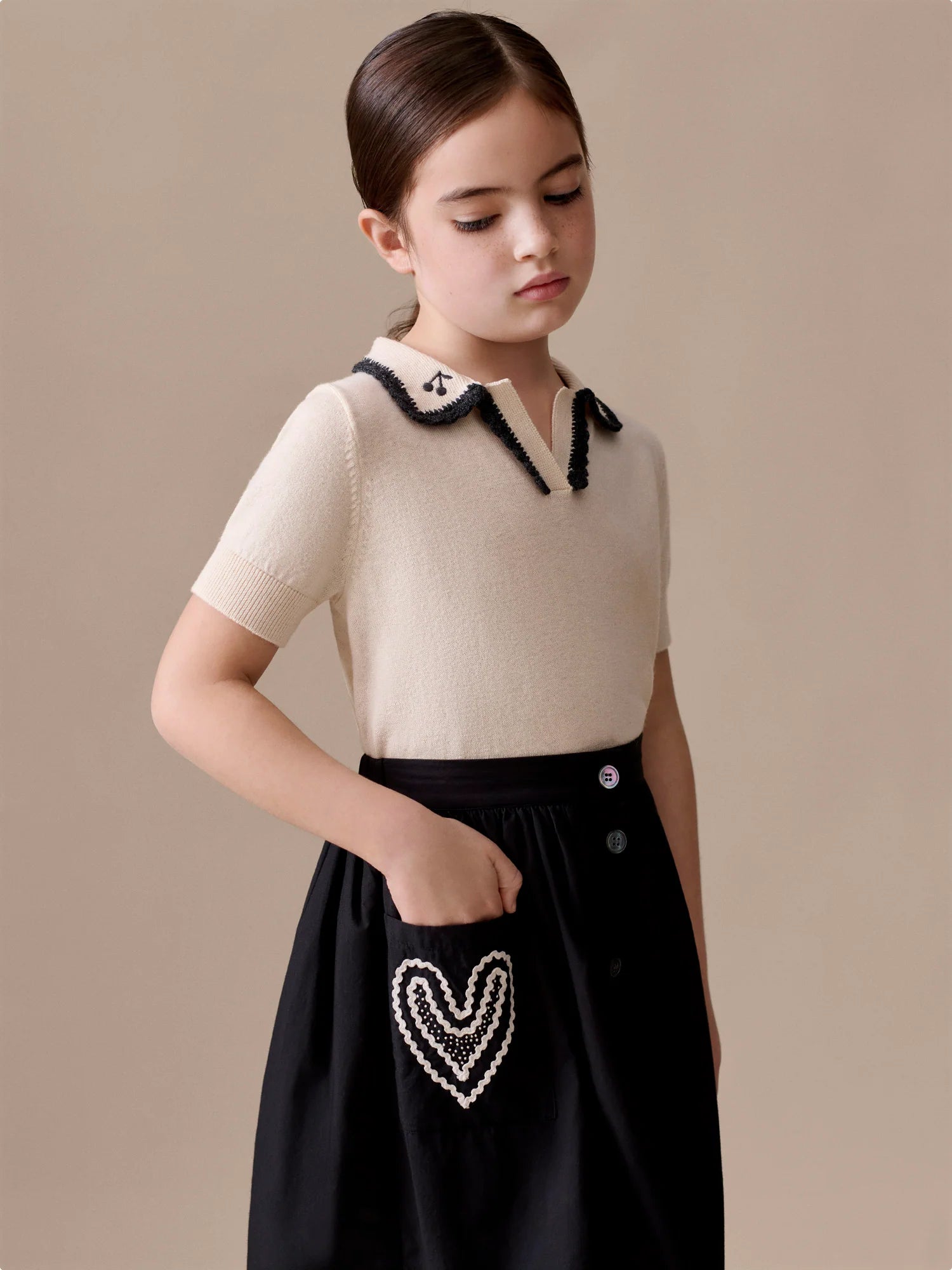 A young girl with dark hair in a low bun wears the Bonpoint Girls' Lisette Sweater with Hand-Embroidered Collar and a black skirt featuring a heart-shaped design on the pocket.