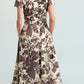 A person stands facing away, wearing the Cara Cara Sawyer Dress—a cream poplin midi dress with a brown floral print and short sleeves—paired with black boots. Their hands are tucked into the dress’s pockets.