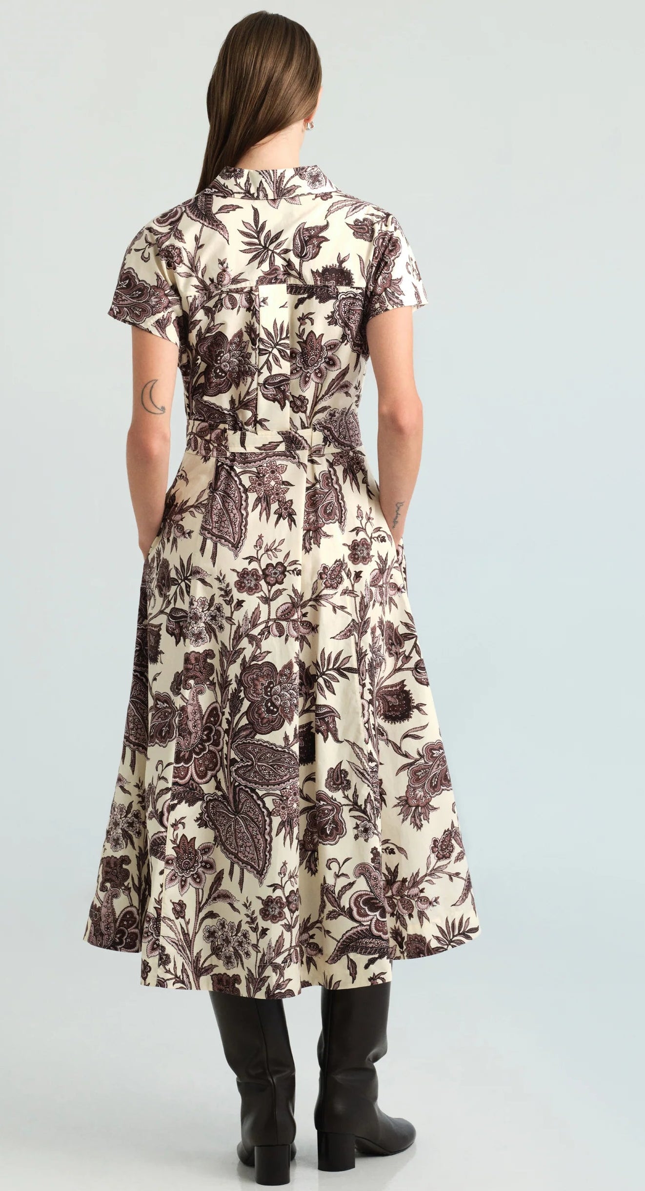A person stands facing away, wearing the Cara Cara Sawyer Dress—a cream poplin midi dress with a brown floral print and short sleeves—paired with black boots. Their hands are tucked into the dress’s pockets.