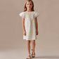 A young girl stands against a beige background, wearing the Bonpoint Girls' Smocked Salomé Dress with a light green cherry print and brown sandals.