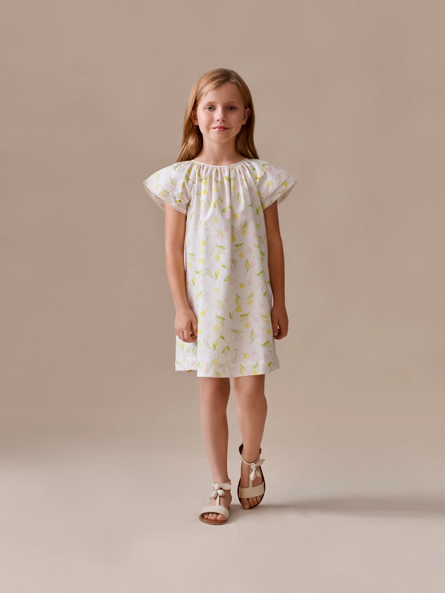 A young girl stands against a beige background, wearing the Bonpoint Girls' Smocked Salomé Dress with a light green cherry print and brown sandals.
