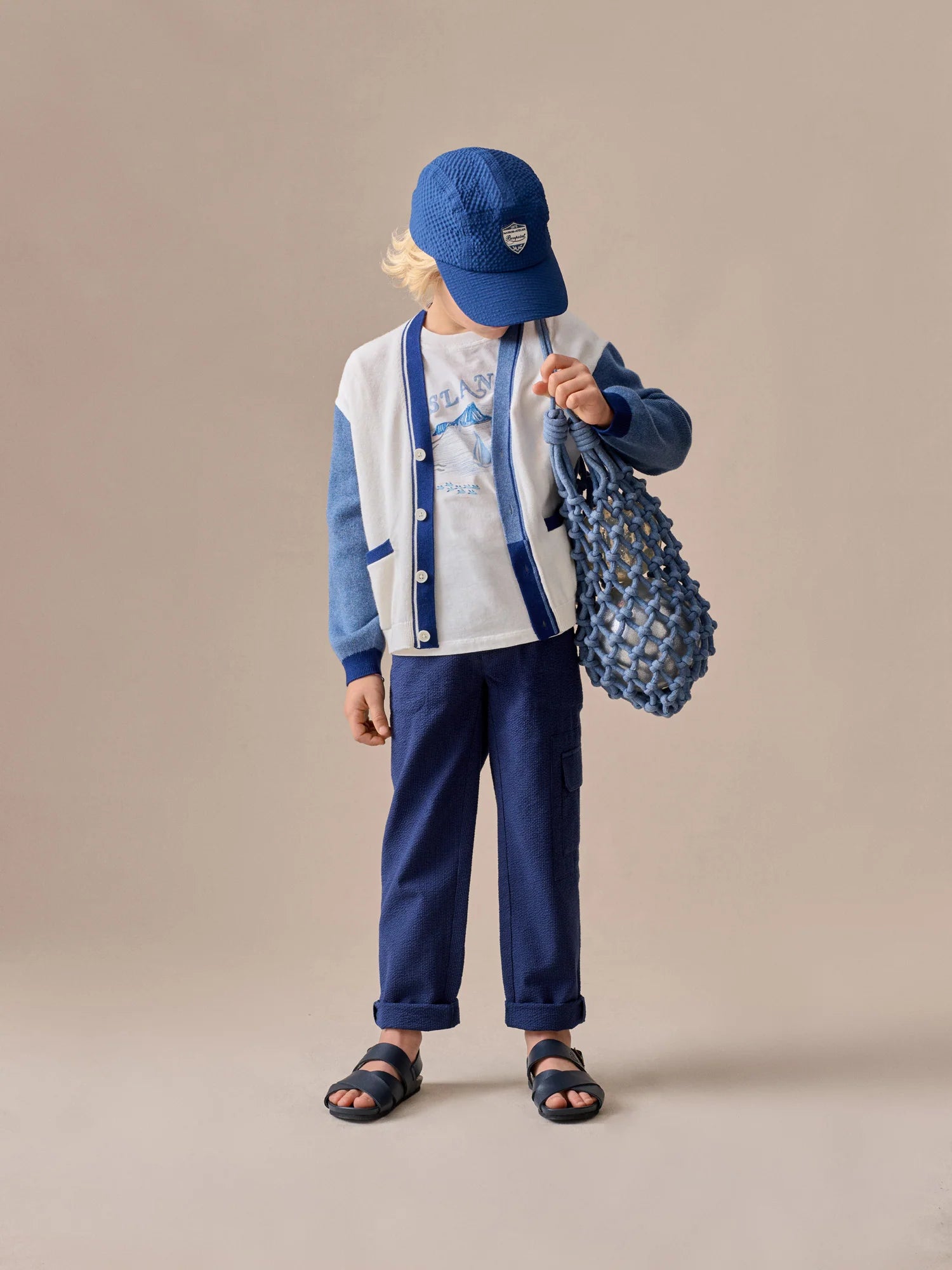 A young boy in a blue cap, white and blue cardigan, graphic tee, Bonpoint Boys' Francis Cargo Trousers, and sandals holds a blue netted bag while standing against a beige background.
