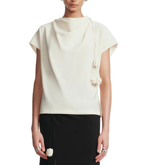 A person is wearing the Lela Rose Drop Flower Cowl Neck Top, a short-sleeve off-white piece with a decorative tie, styled with a black skirt and silver rings on both hands.