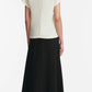A person stands facing away, wearing the Lela Rose Drop Flower Cowl Neck Top in white, paired with a long black skirt and black heeled sandals against a plain background.