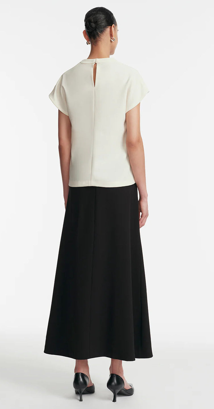 A person stands facing away, wearing the Lela Rose Drop Flower Cowl Neck Top in white, paired with a long black skirt and black heeled sandals against a plain background.