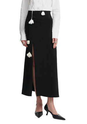 A person wears a white long-sleeve shirt with floral ties, paired with the Lela Rose Drop Flower Ruched Skirt—a black high-waisted A-line midi skirt with a front slit—and black pointed-toe heels.