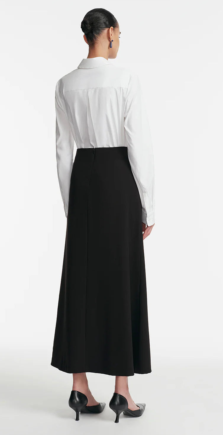 A woman faces away in a white long-sleeve shirt tucked into the Lela Rose Drop Flower Ruched Skirt, featuring an elegant high-waisted A-line silhouette, paired with black heels, set against a plain white background.