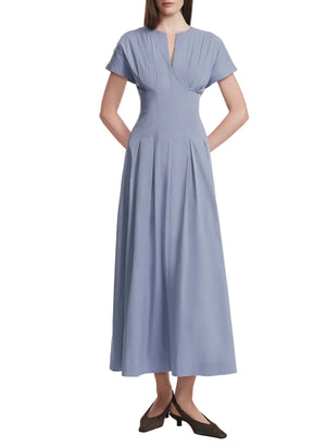 A woman wears the Lela Rose Smocked Back Dress in light blue cotton poplin, featuring a pleat-tailored bodice, V-neckline, ankle-length pleated skirt, and brown shoes.