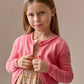 A young girl with long light brown hair wears the Bonpoint Girls' Scalloped Round Neck Larita Cardigan over a patterned dress, standing against a plain beige background.