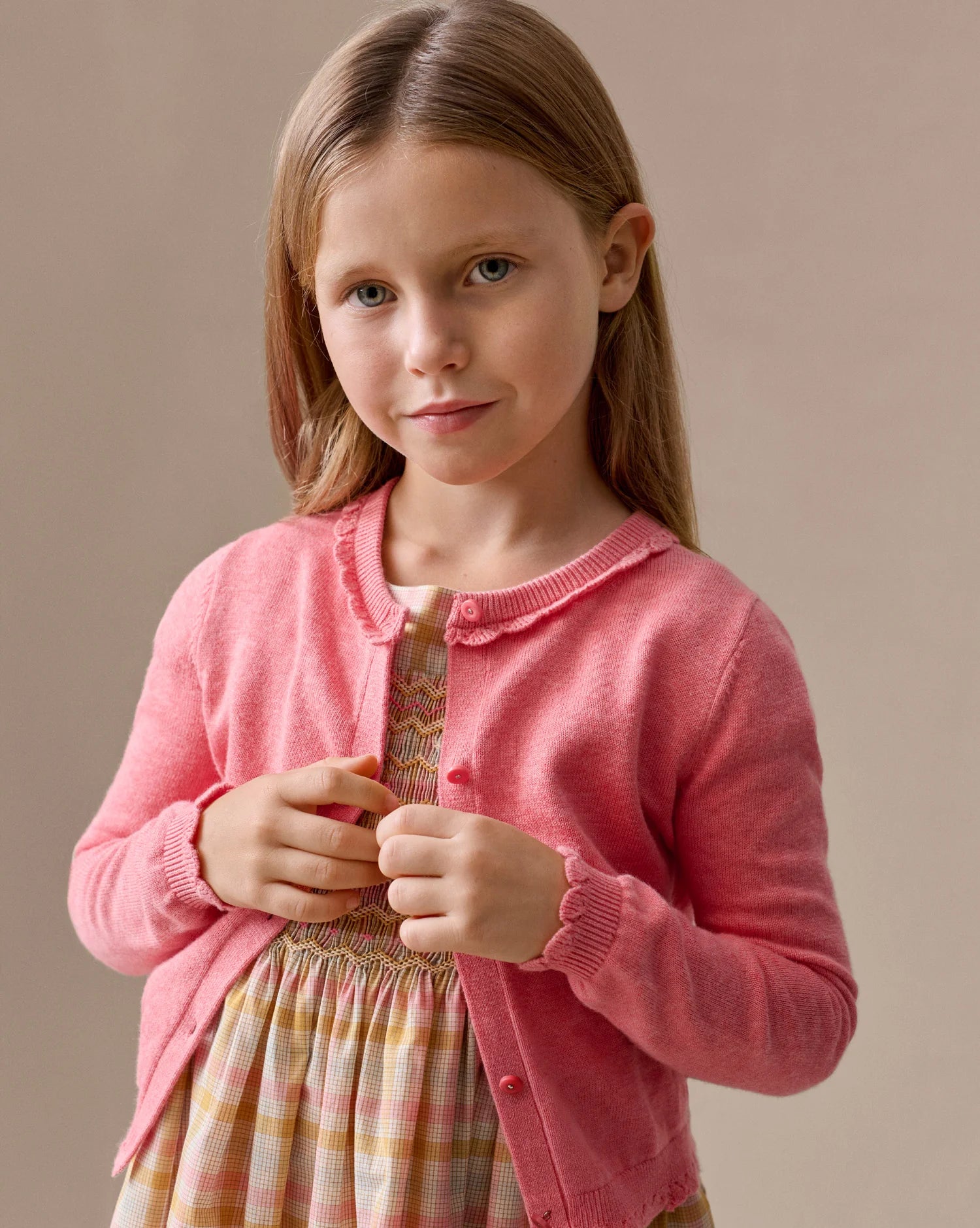 A young girl with long light brown hair wears the Bonpoint Girls' Scalloped Round Neck Larita Cardigan over a patterned dress, standing against a plain beige background.