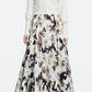 A person stands against a plain white background wearing a Lela Rose Cropped Dolman Cardigan over a long-sleeve white blouse, paired with a high-waisted, full-length abstract-patterned skirt and black heels.