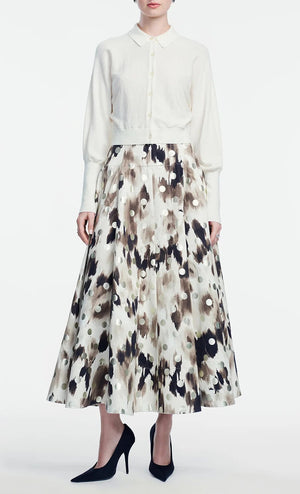 A person stands against a plain white background wearing a Lela Rose Cropped Dolman Cardigan over a long-sleeve white blouse, paired with a high-waisted, full-length abstract-patterned skirt and black heels.