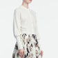 A woman is wearing a cream button-up blouse with a collar and long sleeves, paired with a high-waisted, patterned skirt in beige, brown, and black tones. Her outfit is layered with the Lela Rose Cropped Dolman Cardigan, adding an effortless touch of comfort and style.