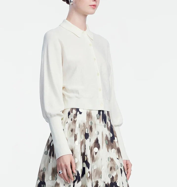 A woman is wearing a cream button-up blouse with a collar and long sleeves, paired with a high-waisted, patterned skirt in beige, brown, and black tones. Her outfit is layered with the Lela Rose Cropped Dolman Cardigan, adding an effortless touch of comfort and style.