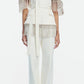 A person wearing the Lela Rose Stretch Cotton Molly Pant, styled with a white tailored pantsuit crafted from stretch cotton, featuring a belted top with fringe details on the sleeves and waist and paired with high-rise straight leg pants for a modern, polished look.